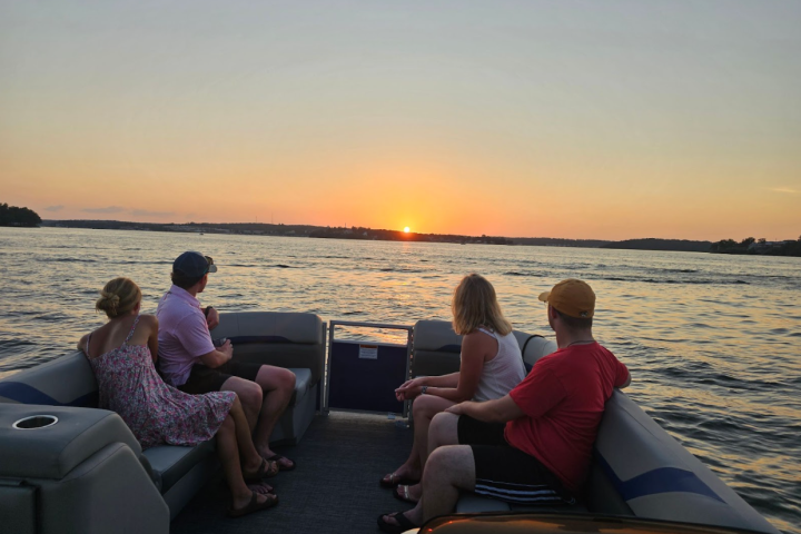 Four people sit on a boat, watching a sunset over a calm lake.