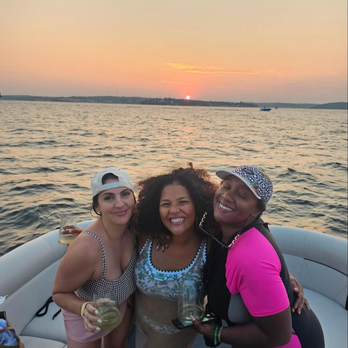 Three people on a boat smiling at sunset, holding drinks on a calm water body.