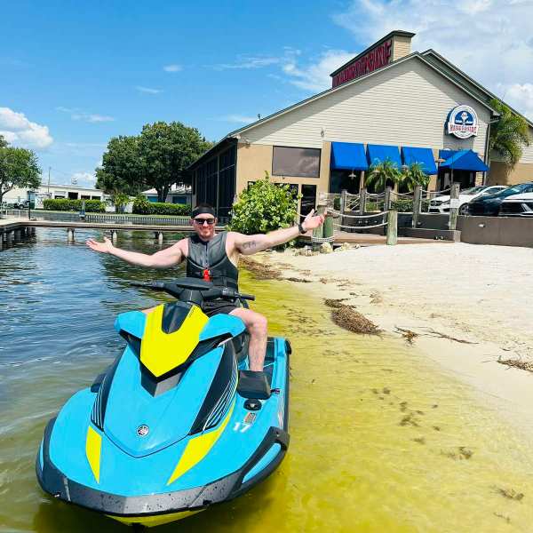 Person on a blue jet ski in shallow water near a waterfront restaurant with a sandy shore.
