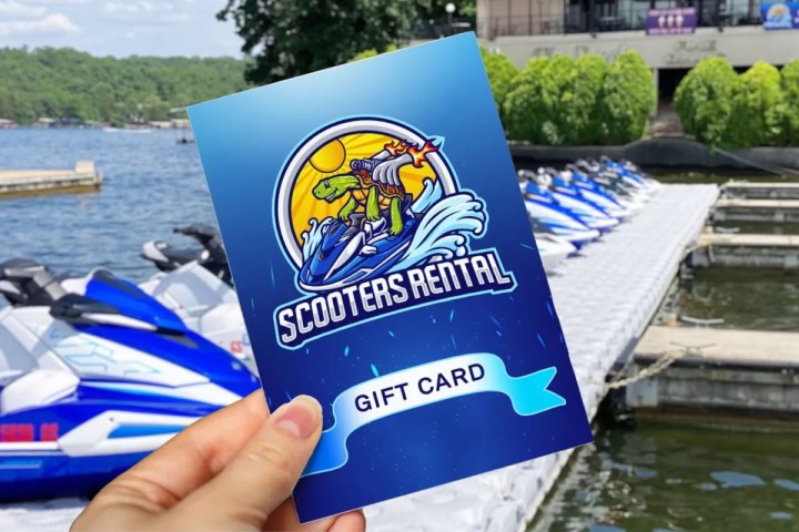 Hand holds Scooters Rental gift card near docked jet skis on a lake.