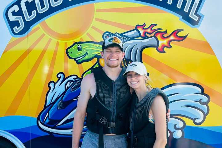Two people in life vests smiling in front of a colorful 'Scooters Rental' sign.