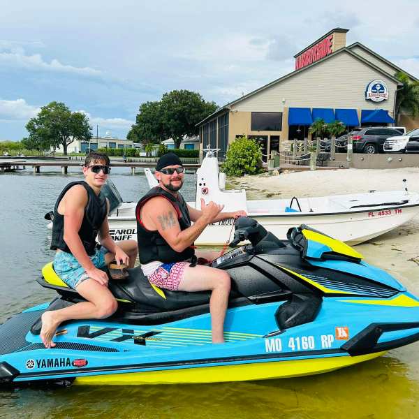 Two people on a jet ski near a lakeside restaurant with parked cars.