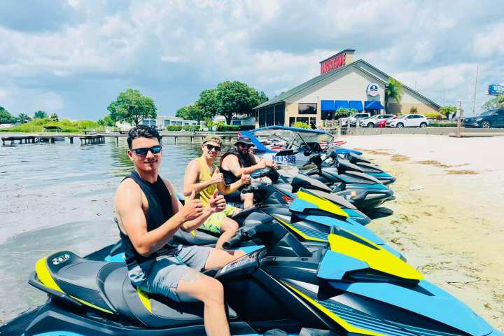 Three people on jet skis by a sandy shore with a restaurant in the background.