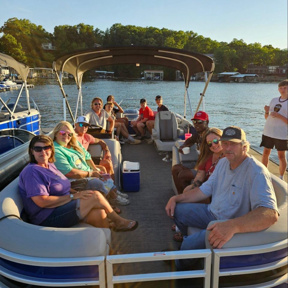 Group of people sitting on a boat with a canopy, on a lake, surrounded by trees.