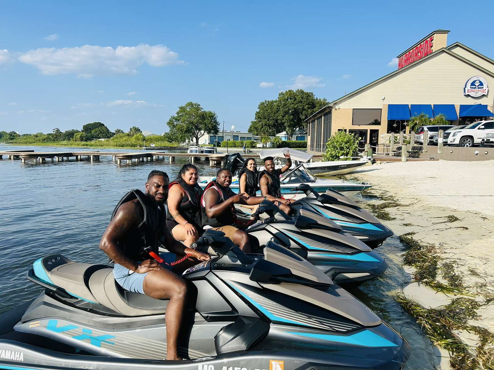 Four people on jet skis by a dock and building on a sunny day.