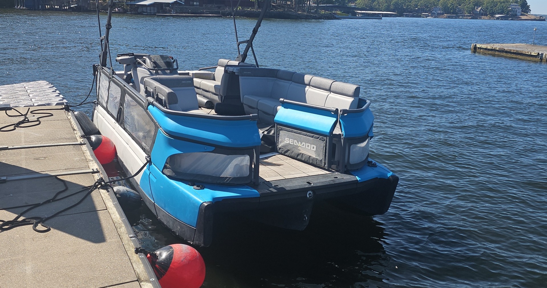 Blue and black boat docked on a sunny lake with buoys and seating.